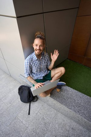 Photo for Informal guy with a backpack and a laptop sits on a granite curb near a modern building - Royalty Free Image