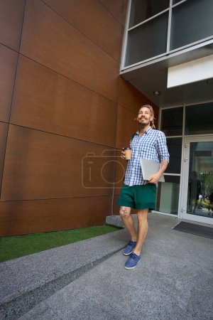 Photo for Young guy is standing on a granite ramp, he has a laptop and a glass of coffee - Royalty Free Image