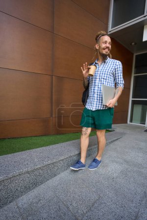 Foto de El joven freelancer se encuentra a la entrada de un moderno edificio de oficinas, tiene una mochila y un portátil - Imagen libre de derechos