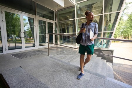 Foto de Freelancer se encuentra a la entrada de un moderno edificio de oficinas, tiene una mochila y un portátil - Imagen libre de derechos