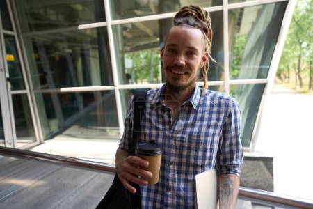 Foto de El tipo informal está parado en un edificio moderno, tiene un portátil y un vaso de café - Imagen libre de derechos