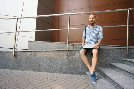 Foto de El tipo informal está sentado en una rampa cerca de un edificio moderno, tiene un portátil y un vaso de café - Imagen libre de derechos
