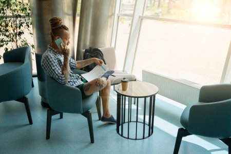 Foto de Hombre informal estudiando documentos de trabajo y hablando por teléfono en el espacio de coworking, él está sentado en sillón por la vista - Imagen libre de derechos