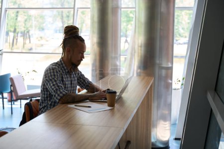 Foto de El hombre informal trabaja en una computadora portátil en una mesa grande, junto a café y papeles de trabajo - Imagen libre de derechos