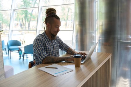 Téléchargez les photos : Informel gars travaille sur un ordinateur portable à une grande table, à côté de café et de documents de travail - en image libre de droit