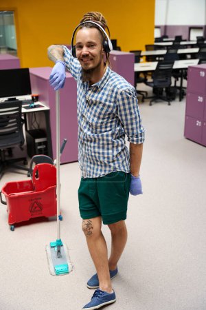 Photo for Happy informal standing in the middle of a coworking space with a mop, he cleans the office space - Royalty Free Image