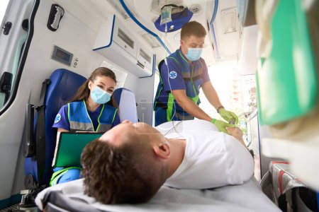 Téléchargez les photos : Les ambulanciers transportent un patient sous goutte à goutte dans une ambulance, une infirmière remplit un questionnaire pour le patient - en image libre de droit
