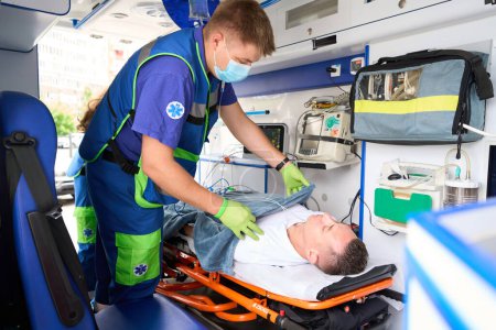 Téléchargez les photos : Homme en gants de protection couvre un patient avec une couverture souple dans une ambulance, le patient se trouve sur une civière spéciale - en image libre de droit