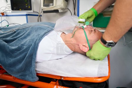 Foto de Hombre con guantes protectores pone una máscara de oxígeno en el paciente en una ambulancia, el paciente se acuesta en una camilla especial - Imagen libre de derechos