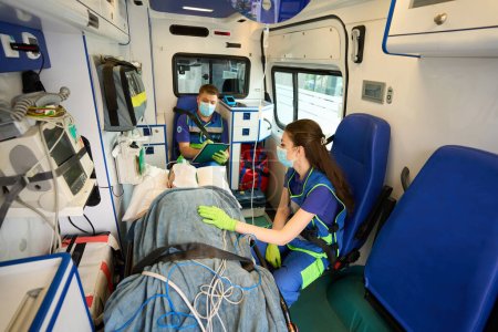 Foto de Médicos del equipo móvil transportan al paciente en máscara de oxígeno en una ambulancia, el médico rellena el cuestionario de pacientes - Imagen libre de derechos