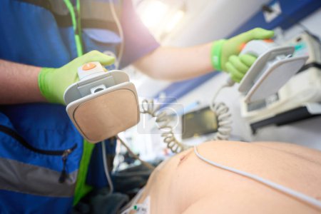 Photo for Employee of a mobile team with defibrillator electrodes in his hands performs resuscitation of a patient in an ambulance - Royalty Free Image