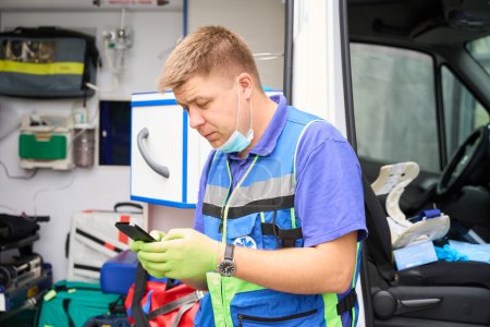 Foto de Médico sostiene un teléfono móvil en sus manos, un hombre está junto a una ambulancia - Imagen libre de derechos