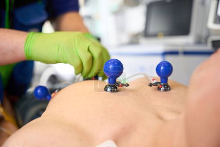 Foto de Médico conecta electrodos en ventosas al torso de un hombre en una ambulancia para tomar un cardiograma - Imagen libre de derechos