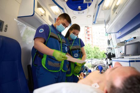 Foto de Un equipo de paramédicos le hace un cardiograma a un hombre en una ambulancia, el paciente yace bajo un goteo - Imagen libre de derechos