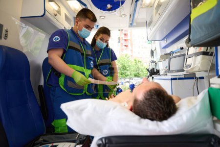 Foto de Los médicos hacen un cardiograma a un hombre en una ambulancia moderna, el paciente se acuesta en una camilla especial - Imagen libre de derechos