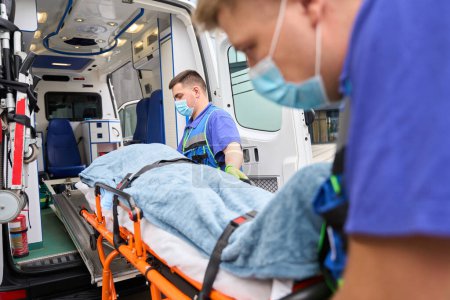 Photo for Young colleagues paramedics unload a gurney with a victim from an ambulance, in the car there is modern equipment - Royalty Free Image