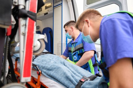 Foto de Médicos experimentados en uniforme descargan una camilla con un paciente de una ambulancia, en el coche hay equipos modernos - Imagen libre de derechos