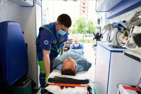 Téléchargez les photos : Les ambulanciers en uniforme déchargent une civière avec un patient d'une ambulance, équipement moderne dans la voiture - en image libre de droit