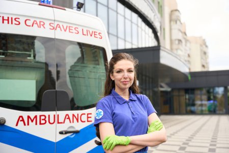 Photo for Female medic stands on the street near an ambulance, she has a medical emblem on her sleeve - Royalty Free Image