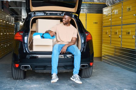 Téléchargez les photos : Mignon Afro-Américain se repose dans le coffre d'une voiture, il a apporté des choses pour le stockage au service - en image libre de droit