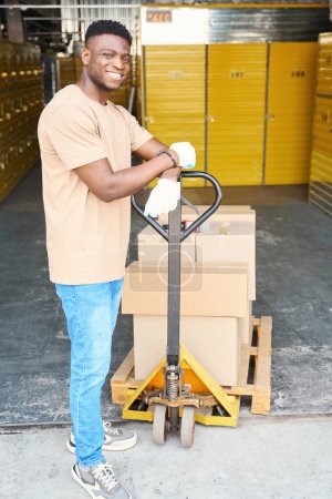 Foto de Joven hombre afroamericano en un almacén con un carro de carga, en el carro hay cajas de cartón con cosas - Imagen libre de derechos