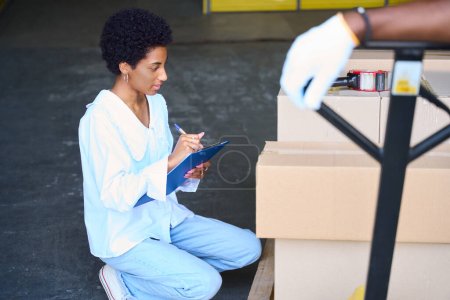Téléchargez les photos : Femme gestionnaire à l'entrepôt fait un inventaire des boîtes en carton en cours de dépôt, à côté d'un homme avec un chariot de fret - en image libre de droit