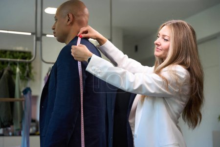 Photo for Focused female tailor measuring adult male client half-shoulder width with tape measure - Royalty Free Image