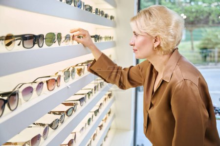 Photo for Client of an optical salon in a brown blouse chooses sunglasses, there is a wide selection of glasses on display - Royalty Free Image