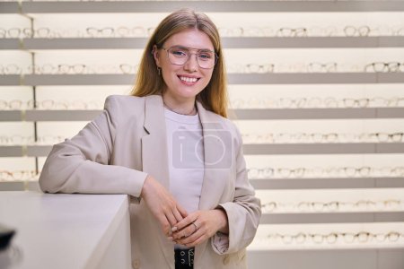 Foto de Mujer joven en puestos de gafas en el mostrador en el salón de óptica, hay una amplia selección de gafas en escaparate - Imagen libre de derechos
