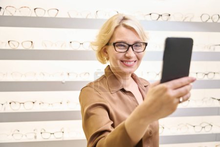 Photo for Woman in a brown blouse takes a selfie wearing new glasses, there is a wide selection of glasses on display - Royalty Free Image