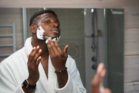 Photo for Young male applies shaving foam to his cheeks, and chin as he goes through his morning routine in the bathroom - Royalty Free Image