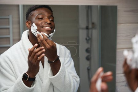 Photo for Cute male applies shaving foam to his cheeks, while doing his morning routine in the bathroom - Royalty Free Image