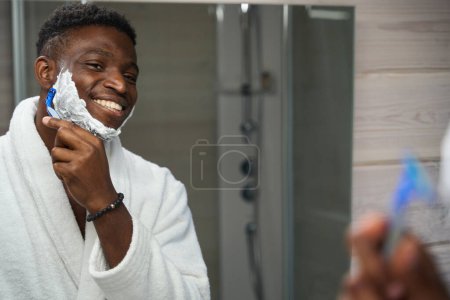Photo for African American guy shaves in front of the mirror in the bathroom, he is wearing a terry robe - Royalty Free Image