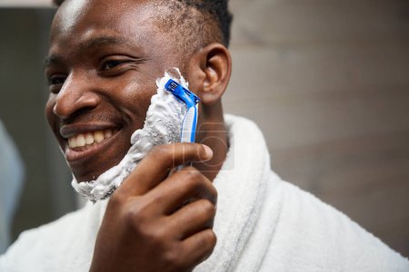 Téléchargez les photos : Afro-américain mec se rase avec un rasoir jetable, il utilise de la mousse à raser - en image libre de droit