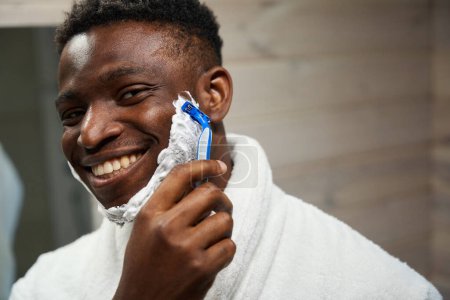 Photo for Smiling African American male shaves with a disposable razor, he uses shaving foam - Royalty Free Image