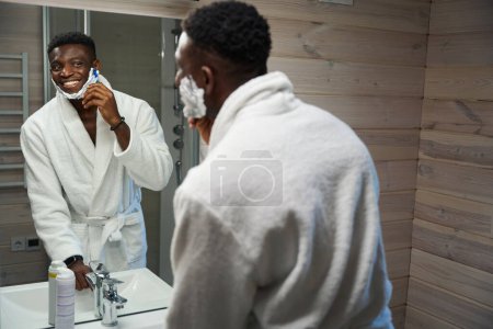 Photo for African American man shaves in front of a large mirror in the bathroom, he is wearing a terry robe - Royalty Free Image