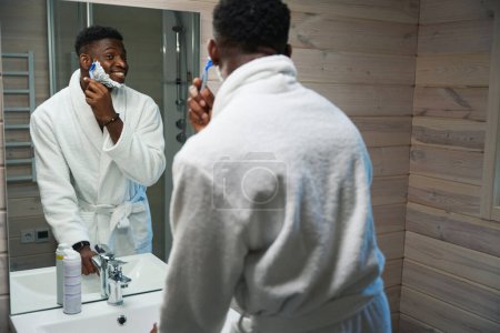 Téléchargez les photos : Jeune homme se rase devant un grand miroir dans la salle de bain, il porte une robe éponge - en image libre de droit