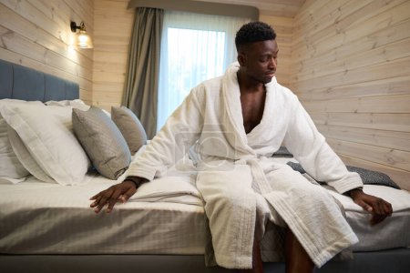 Téléchargez les photos : Homme aux cheveux bouclés assis sur le bord du lit dans une chambre confortable, il porte un peignoir éponge doux - en image libre de droit