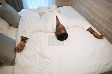 Foto de Feliz hombre se encuentra en la cama en un acogedor dormitorio, él está usando una túnica de rizo suave - Imagen libre de derechos
