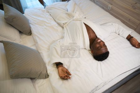 Foto de Sonriente hombre se encuentra en una cama grande en un acogedor dormitorio, que lleva una túnica de rizo suave - Imagen libre de derechos