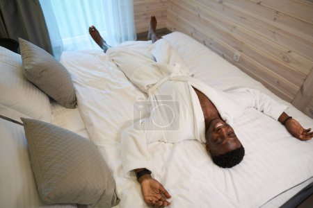 Foto de Sonriente hombre afroamericano se encuentra al otro lado de la cama en un acogedor dormitorio, lleva una suave bata de baño - Imagen libre de derechos