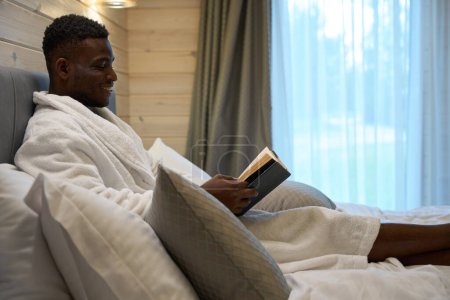 Photo for Male enjoys morning reading in a cozy bedroom, he is located on a large bed - Royalty Free Image