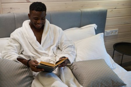 Foto de Hombre afroamericano disfruta de la lectura de la mañana en un acogedor dormitorio, él está sentado en una cama grande - Imagen libre de derechos