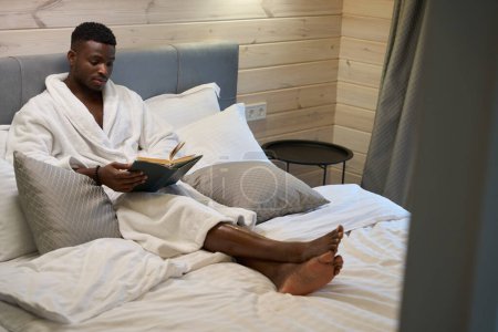 Foto de Hombre afroamericano guapo está leyendo un libro en un acogedor dormitorio, él está sentado en una cama grande - Imagen libre de derechos