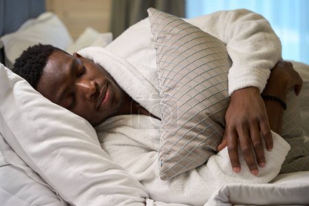 Photo for African American guy in a terry robe sleeps sweetly on a large bed, in a modern bedroom interior - Royalty Free Image
