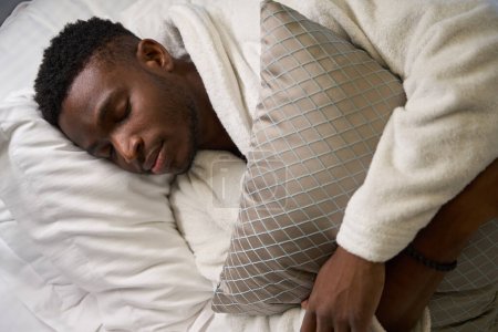 Téléchargez les photos : Afro-Américain en peignoir éponge dort doucement sur un grand lit, dans son sommeil, il étreint un oreiller - en image libre de droit