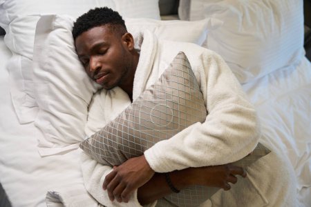 Foto de El tipo de pelo rizado con una bata de felpa duerme dulcemente en una cama grande, mientras duerme abrazó una almohada - Imagen libre de derechos