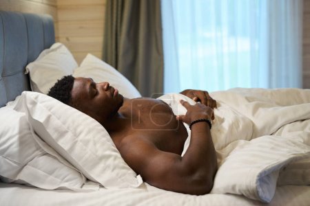 Foto de Chico afroamericano con un torso desnudo duerme dulcemente en una cama grande, amanecer fuera de la ventana - Imagen libre de derechos