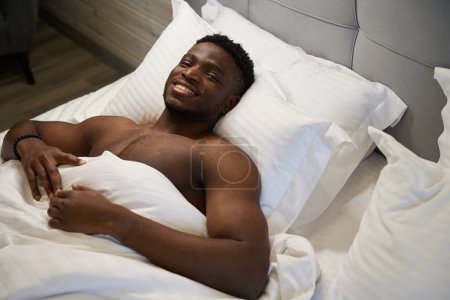 Photo for African American man with a naked torso enjoys morning relaxation in a soft bed, he is located on soft pillows - Royalty Free Image