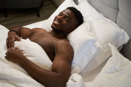 Photo for Smiling man with a naked torso enjoys morning relaxation in a soft bed, he is sitting on soft pillows - Royalty Free Image